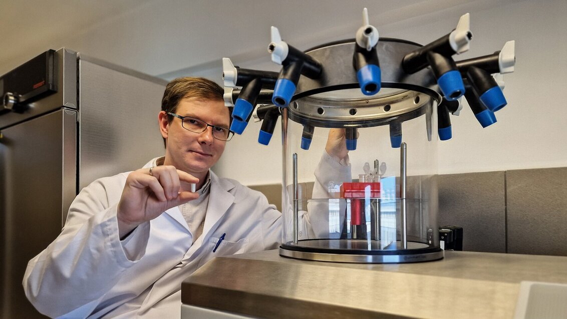 Na zdjęciu dr inż. Michał Dziadek w fartuchu laboratoryjnym przy specjalistycznym sprzęcie laboratoryjnym. W prawej dłoni trzyma małą szklaną próbkę.