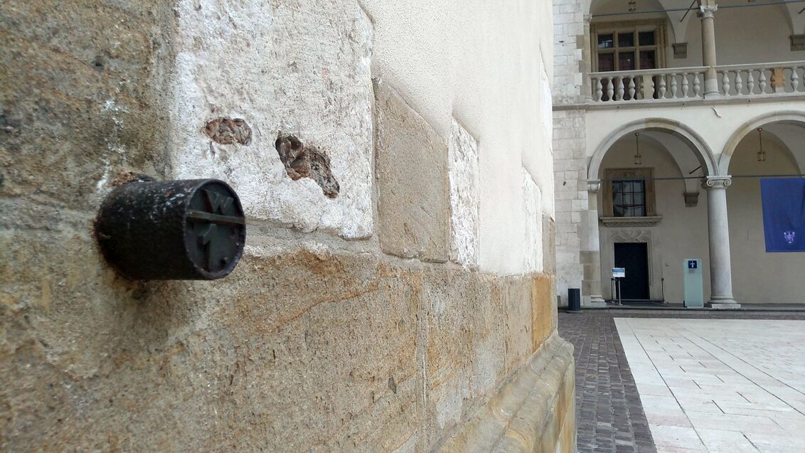 A benchmark on a wall of a historic building with arcades in the background.