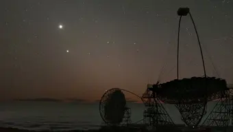 Nocne niebo pełne gwiazd nad linią brzegową stworzoną przez chmury, na pierwszym planie widoczne duże konstrukcje radioteleskopów w formie ażurowych anten.