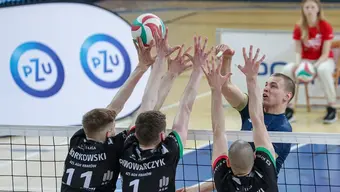 Mecz siatkówki. Z jednej strony siatki trzech zawodników, z drugiej - jeden. Wszyscy wystawiają ręce w stronę piłki, która jest nad siatką. Zawodników widać od pasa w górę. Trzech zawodników na pierwszym planie widzimy od tyłu. W tle widoczne logo sponsora zawodów PZU.