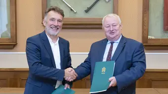 Two man in elegant attire shaking hands while holding green folders and smiling. 