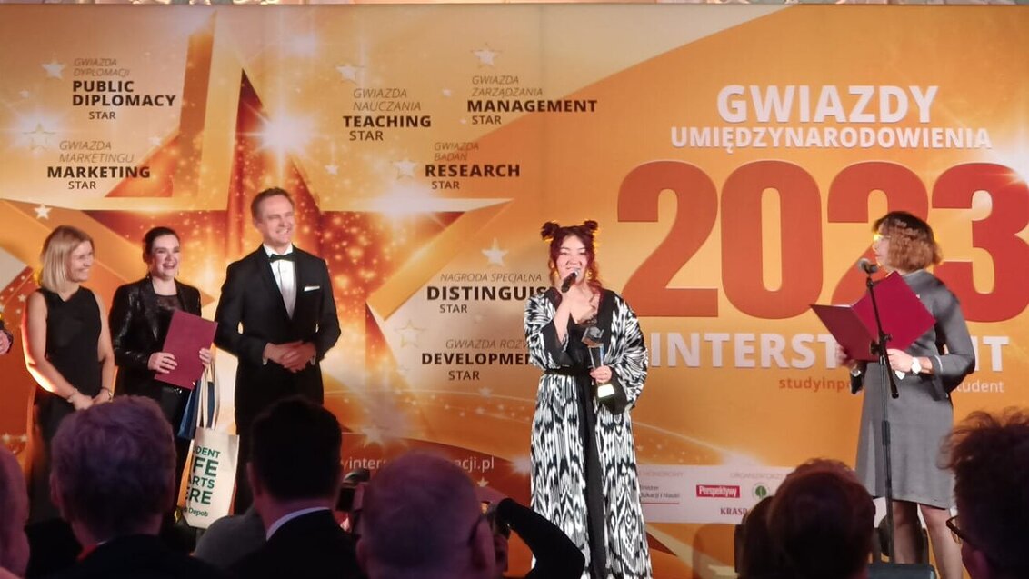 Zdjęcie z rozdania nagród. Laureatka stoi na scenie, przemawiając do mikrofonu, obok niej organizatorzy konkursu.