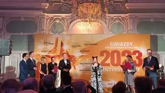 Photo from the presentation of the awards gala. The laureate stands on stage and speaks to the microphone. Besides her, there are the organisers of the competition.