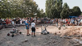 A group of people gathered around Martian rovers on a rocky surface outdoors. a large number of spectators is surrounding the track while the individuals are operating the robots.
