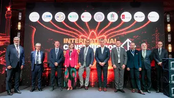 Group of formally dressed attendees posing on a stage at an international construction conference, with logos displayed on a large screen behind them.