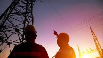 Na zdjęciu dwóch energetyków w kaskach ochronnych przy słupach wysokiego napięcia.