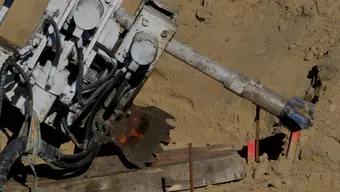 Zdjęcie ciężkiej maszyny budowlanej podczas pracy w terenie – widoczne przewody hydrauliczne i metalowe elementy, w tym duże ramię cylindryczne zagłębiające się w piaszczyste podłoże.