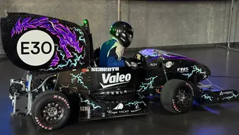 A sleek black racing car with bold purple lightning graphics, sponsor logos, and a fully geared driver inside. It’s showcased indoors with a screen displaying a digital rendering of a similar car. 