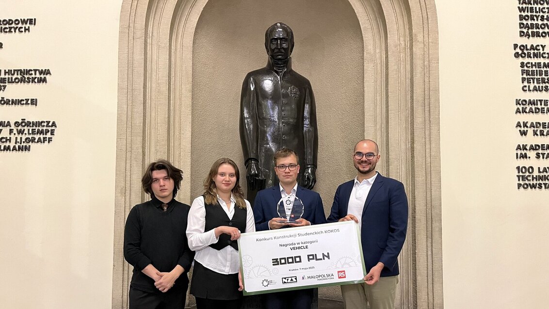 Na zdjęciu cztery młode osoby trzymające wspólnie planszę z nazwą konkursu i wymieniona kwotą nagrody (3000 zł). Za osobami widoczny posąg Stanisława Staszica. Zdjęcie wykonane na schodach w holu reprezentacyjnym AGH.