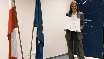 Photo of the laureate. She holds a commemorative diploma in her hands and behind her is a POLSA promotional roll-up banner. Next to her, there are flags of Poland and the European Union.