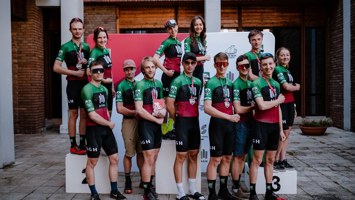 A group photo of the mountain biker team from the AGH University (13 people in total). They are wearing sports clothes in university colours. They all have medals hanging on their necks. One of the contestants is holding a commemorative statuette.