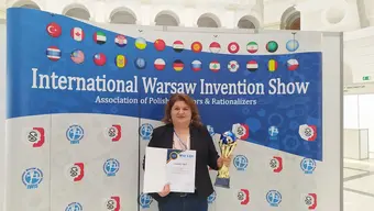 Image of Dr Ewa Knapik standing in front of a photo wall with the IWIS and parter logotypes and flags of representatives, holding the grand prix award