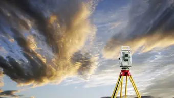 Na zdjęciu geodezyjny przyrząd pomiarowy na tle zachmurzonego nieba.