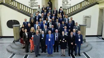 Grupa kilkudziesięciu uczestników konferencji pozuje do wspólnego zdjęcia na reprezentacyjnej klatce schodowej. W tle znajduje się rzeźba oraz ściany z napisami i datami.