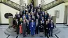 Grupa kilkudziesięciu uczestników konferencji pozuje do wspólnego zdjęcia na reprezentacyjnej klatce schodowej. W tle znajduje się rzeźba oraz ściany z napisami i datami.