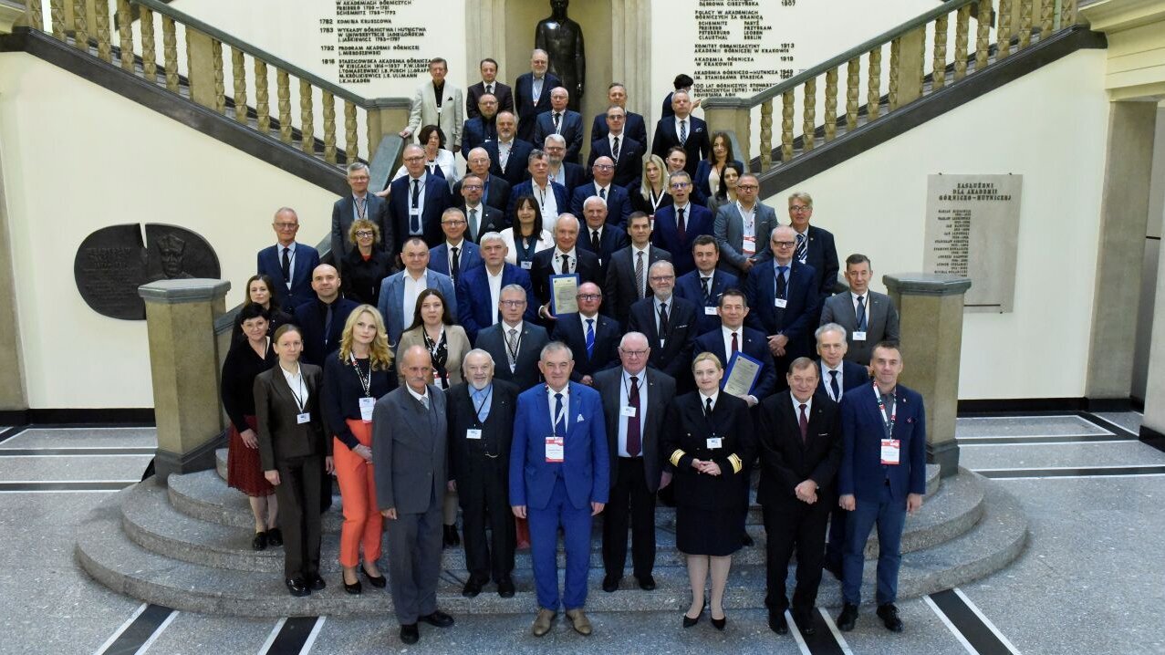 Grupa kilkudziesięciu uczestników konferencji pozuje do wspólnego zdjęcia na reprezentacyjnej klatce schodowej. W tle znajduje się rzeźba oraz ściany z napisami i datami.
