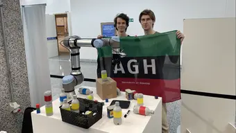 Image of two male studens holding an AGH University banner standing behind a desk with a robotic arm and various objects