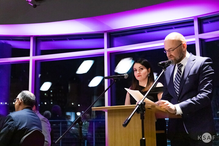 Na zdjęciu mężczyzna stojący przy mównicy z mikrofonem i wygłaszający przemówienie. Po jego prawej stronie stoi kobieta, widocznych jest dwóch mężczyzn siedzących przy stole. W tle fioletowe światło.