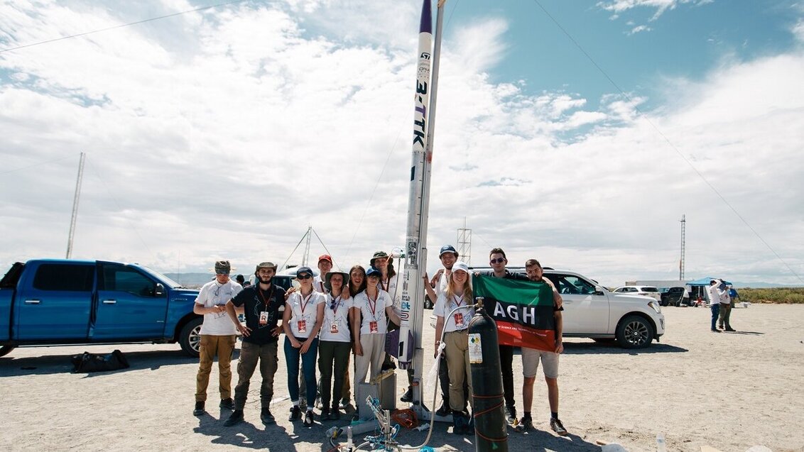 A group of people with a rocket behind them. Cars in the background. Sunny day, blue sky and white, frothy clouds.