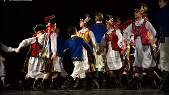 Zdjęcie przedstawia grupę tańczących na scenie dzieci w strojach ludowych.