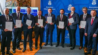 Uczestnicy wydarzenia stoją przed ścianką z logo Prezydenta Miasta Krakowa, trzymając w dłoniach podpisane dokumenty.