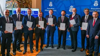 A group of officials standing indoors in front of a blue Krakow city backdrop, each holding a signed document following the signing of a letter of intent.