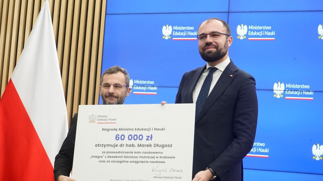 Image of the Minister of Education and Science with Dr Długosz from the AGH University holding the award, a large banner with the prize of PLN 60,000; Polish flag visible on the left and blue background with the Ministry's logotype