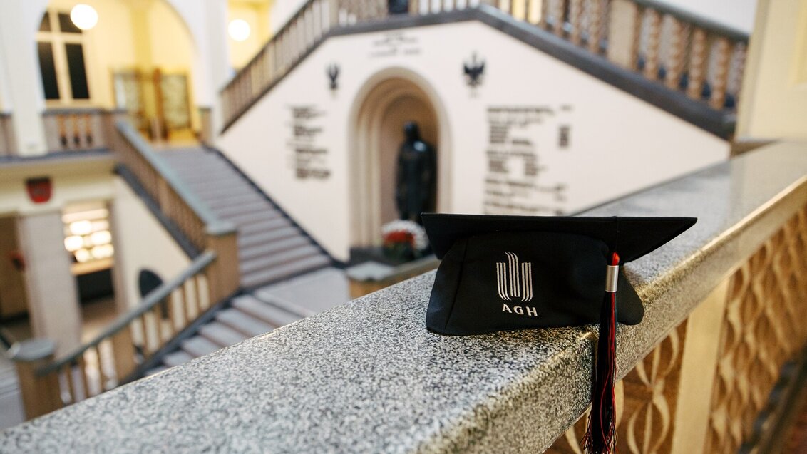 In the foreground, there is a black graduate cap with the university logo embroidered in light beige. There is a tassel attached to the cap, the strings of which are black, red, and green. The cap lies on the railing of a pillared cloister in the Main Building. In the background, a fragment of the main hall of the Main Building is visible as seen from the first floor: stairs, a niche with the statue of Stanisław Staszic, hallways.