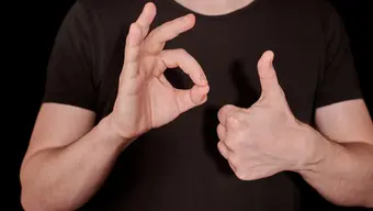 Osoba w czarnej koszulce na czarnym tle wykonuje gesty języka migowego: prawą ręką pokazuje znak 'OK', a lewą ręką gest 'kciuk w górę'.