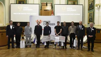 Ceremonia wręczenia nagród Prize Charger w auli AGH, laureaci stoją z symbolicznymi czekami i upominkami przed banerem z logo wydarzenia.