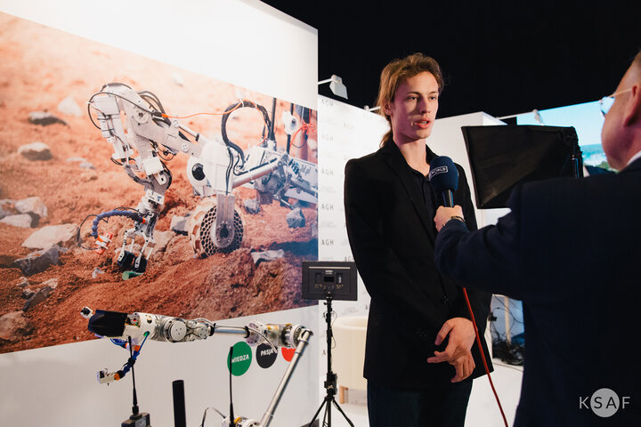 A young man being interviewed by someone holding a microphone. A large poster photo of a Martian rover on the wall behind the man.