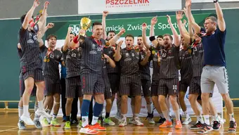The photo shows the winning male AZS AGH handball team. A group of players stands on the field floor. They wear sports clothes. One of them holds a gold cup. All players cheer for their victory with their hands raised. Their mouths are open, screaming. Their faces look very happy.