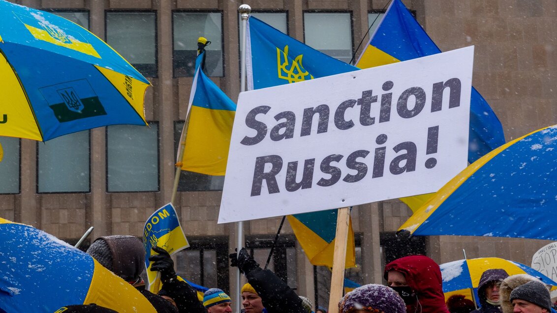 Fotografia przedstawia mężczyzn i kobiety ubranych w zimowe stroje, którzy trzymają flagi państwowe Ukrainy oraz parasole w narodowych barwach ukraińskich. Nad nimi widoczny jest również transparent z napisem „Sanction Russia!”.