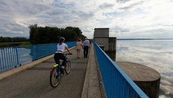 Ludzie przemieszczają się pieszo i rowerem po drodze biegnącej nad wodą i ograniczonej barierkami