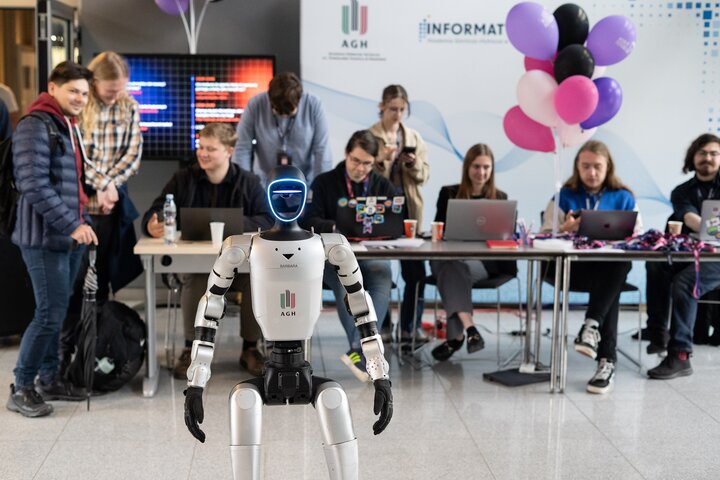 A humanoid robot with the logotype of AGH University on its torso standing in the middle of a room. Behind it, a number of students are sitting at a table.