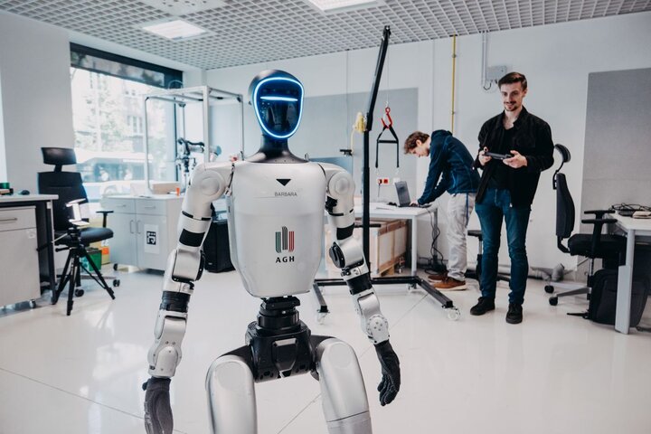 Humanoidalny robot z podświetlaną twarzą stoi w laboratorium. W tle dwóch naukowców pracuje przy sprzęcie i komputerze.