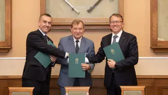Na zdjęciu trzech mężczyzn w garniturach - sygnatariuszy umowy. Stoją obok siebie, uśmiechają się, podają sobie dłonie we wspólnym uścisku. Zdjęcie wykonane w sali konferencyjnej, tuż po podpisaniu dokumentów.