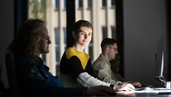 An image of four people sitting at desks in front of computer screens, with one person, a male student, looking directing to the camera.