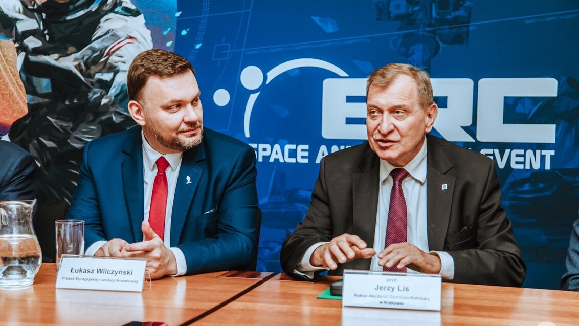 Zdjęcie z konferencji prasowej. Widoczne dwie osoby - Rektor JAGH Jerzy Lis i Łukasz Wilczyński - siedzą obok siebie, za stołem. Za nimi baner zawodów.