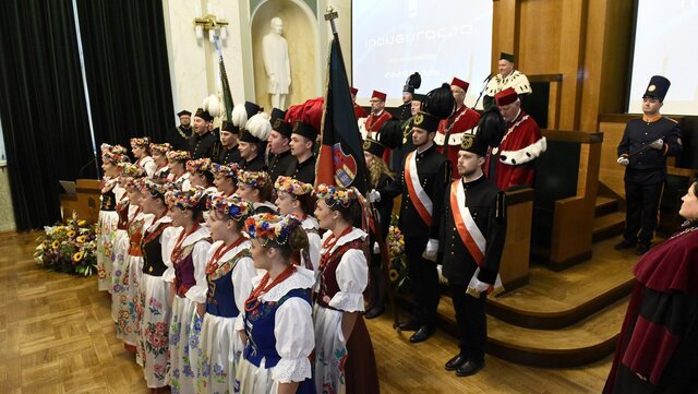 Uroczystość inauguracji roku akademickiego w auli AGH. Na pierwszym planie członkowie i członkinie Zespołu Pieśni i Tańca AGH „Krakus” w strojach ludowych, obok poczet sztandarowy, a w tle przedstawiciele władz uczelni w togach akademickich.