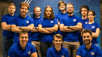 Zdjęcie grupowe członków i członkiń (w sumie 11 osób) Koła Naukowego AGH Lunar Technologies. Wszyscy ubrani w jednakowe niebieskie koszulki polo z małym elementem graficznym w kolorze białym.