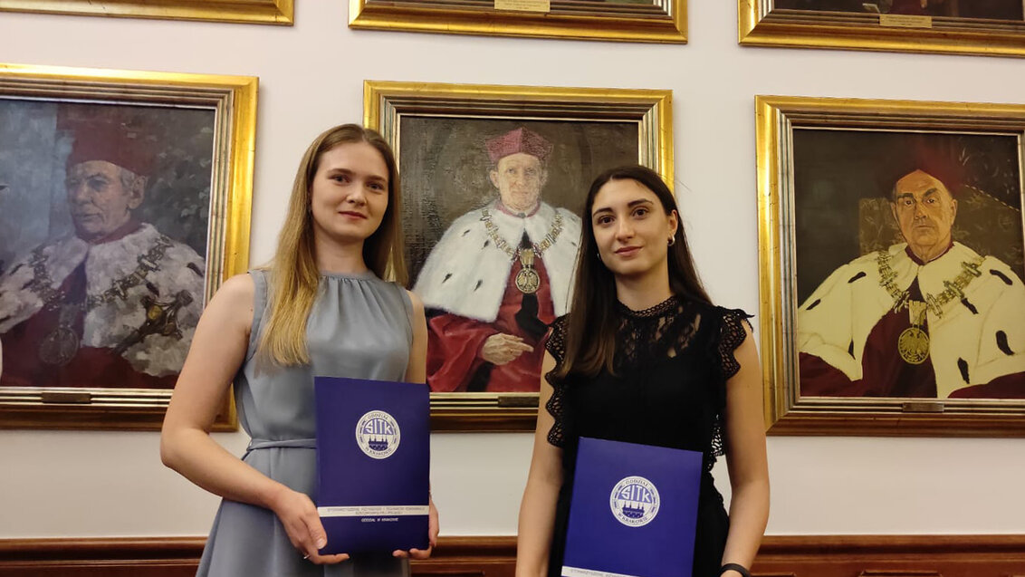 Dwie studentki trzymają w rękach niebieskie teczki z dyplomami. Za nimi ściana z obrazami rektorów. 