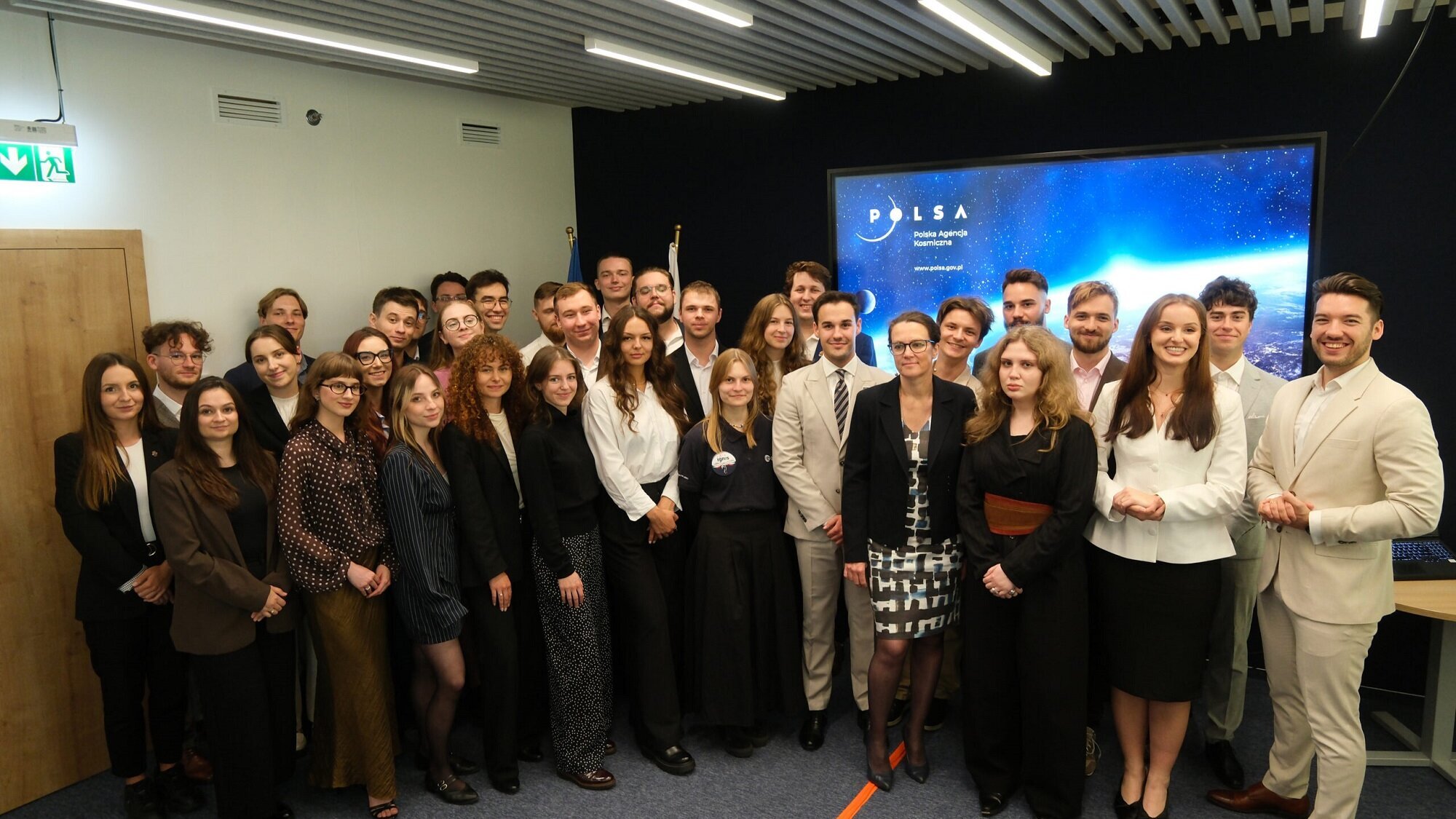 Członkowie i członkinie VII kadencji Rady Studentów przy Prezesie POLSA podczas spotkania inauguracyjnego - pozują wraz z prezes POSLA do wspólnego zdjęcia na tle ekranu z logo Polskiej Agencji Kosmicznej.