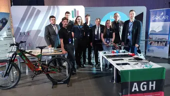 Zdjęcie grupowe na stoisku AGH podczas kongresu Nauka dla Biznesu. Widoczna ekspozycja projektów studenckich (rower elektryczny). Na stole materiały informacyjne i gadżety, w tle banery AGH i kół naukowych.