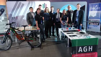 A group of people at an AGH University exhibition stand. An electric bicycle is positioned to the left, and a table on the right-hand side is adorned with the AGH University flag.