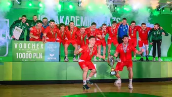 Grupa zawodników w czerwonych strojach sportowych stoi na podium. Na szyjach mają zawieszone medale. Dwóch z nich trzyma duży, pamiątkowy puchar. na twarzach zawodników widoczna duma i radość. W tle zielona ściana z napisem „Liga Akademicka AZS” oraz efekty dymne. Na podium widoczny symboliczny, duży voucher o wartości 10 000 zł na sprzęt sportowy.