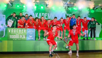 An image of a group of handball players in red uniforms on a podium, cheering and raising a trophy amid green lights and smoke effects, with medals around their necks and a large prize voucher displayed in front.