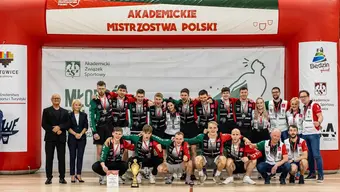 Grupowe zdjęcie zawodników wraz z trenerami. Na szyjach zawodników zawieszone medale, przed nimi na parkiecie stoi pamiątkowy puchar.