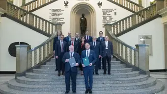 Grupa 12 elegancko ubranych osób stoi na schodach w holu budynku, za nimi w tle posąg Stanisława Staszica