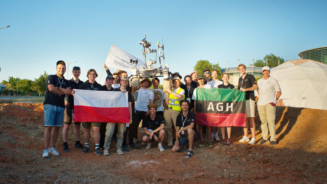 A group photo of the AGH Space Systems team during the Anatolian Rover Challenge 2026. The participants are holding an AGH University flag and a Polish flag. In the centre, several members of the group are lifting the rover above their heads.
